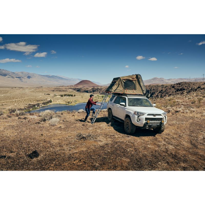 Tuff Stuff Overland Alpha II Hard Shell Rooftop Side Open Tent (2 Person). Man climbing ladder to go into open tent on vehicle